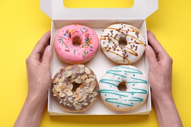 Photo of Woman holding box with tasty glazed donuts on yellow background, top view