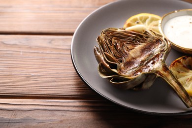 Photo of Tasty grilled artichoke, slices of lemon and sauce on wooden table, closeup. Space for text