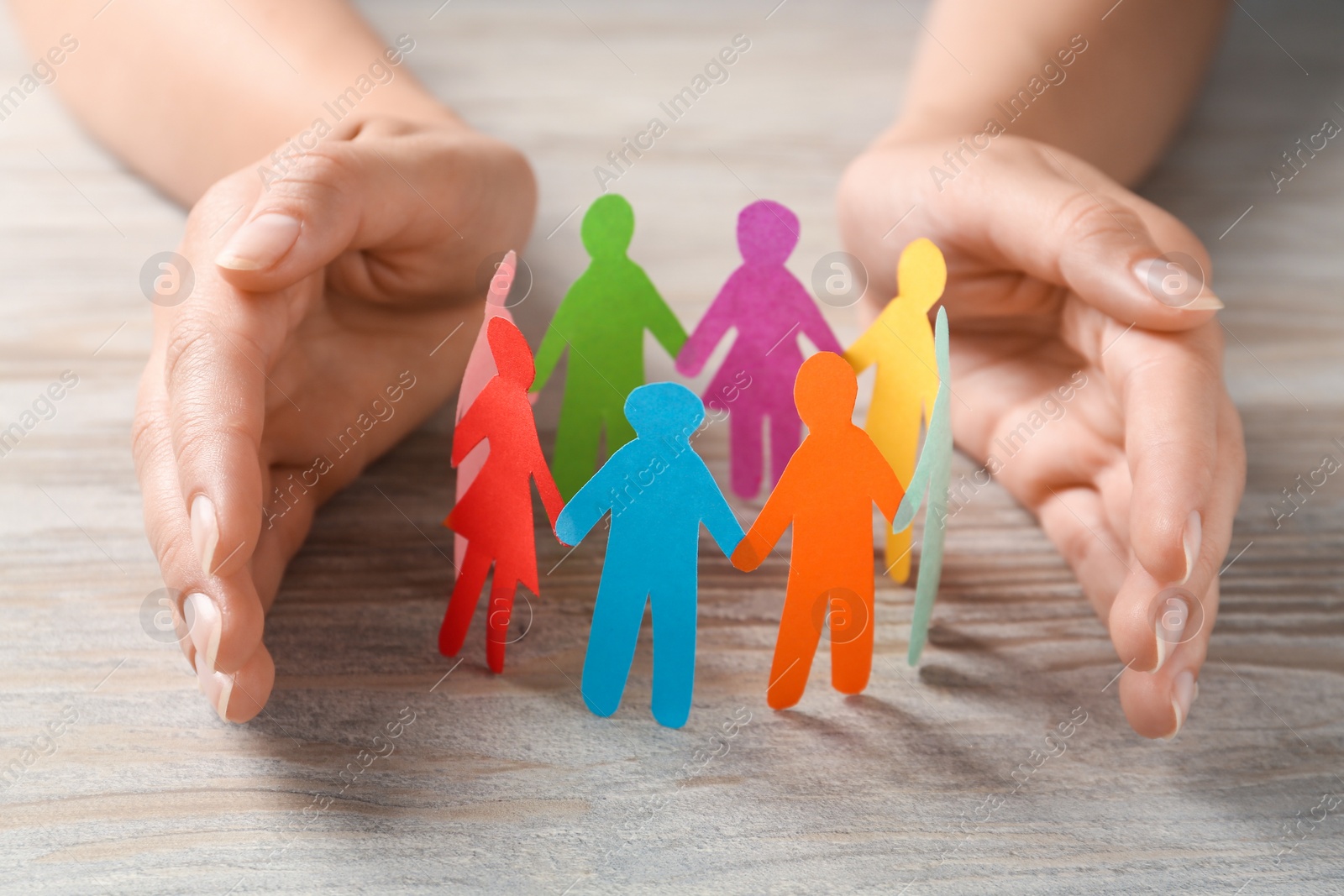 Photo of Woman protecting paper human figures on wooden table, closeup. Diversity and inclusion concept