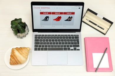 Photo of Online store. Laptop with open website, notebook, pen, croissant and pencil on wooden table, above view