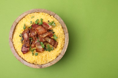 Photo of Cooked cornmeal with bacon and microgreens in bowl on light green table, top view. Space for text