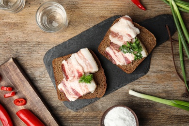 Board with pork fatback sandwiches, shots of vodka, onion and pepper on wooden background, flat lay