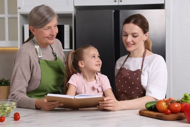 Cute little girl with her mother and grandmother cooking by recipe book in kitchen