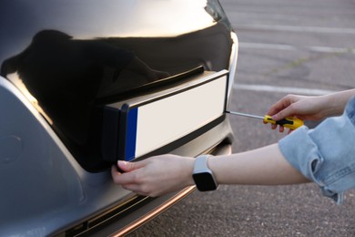 Photo of Woman with screwdriver installing vehicle registration plate to car outdoors, closeup