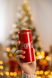 Photo of MYKOLAIV, UKRAINE - January 01, 2021: Woman with can of Coca-Cola against blurred Christmas tree indoors, closeup