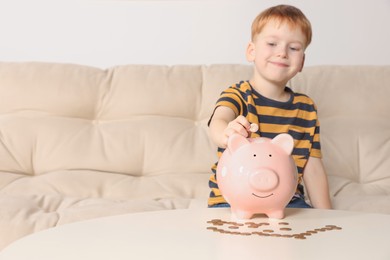Photo of Cute little boy putting coin into piggy bank at table indoors, focus on hand. Space for text