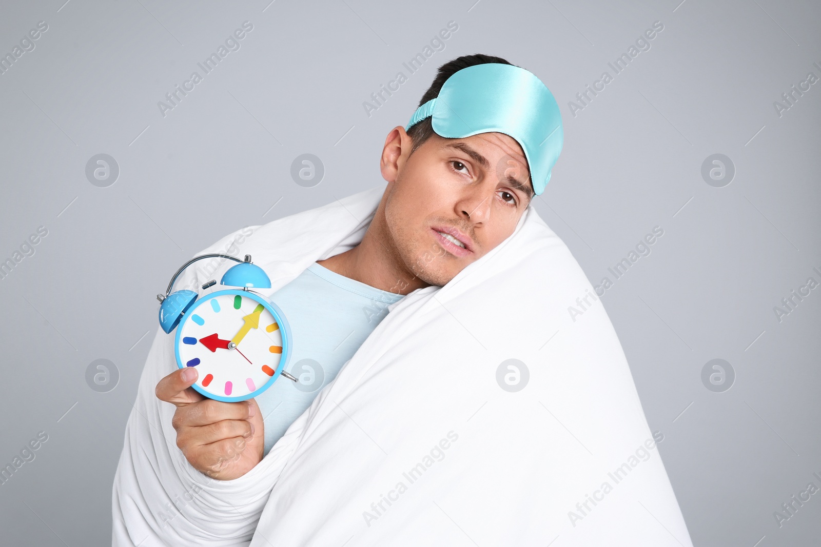 Photo of Man in sleeping mask wrapped with blanket holding alarm clock on grey background