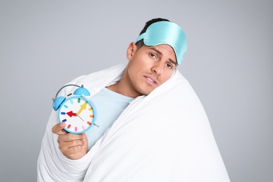 Man in sleeping mask wrapped with blanket holding alarm clock on grey background