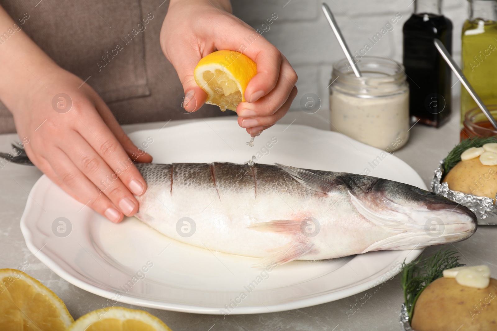 Photo of Woman squeezing lemon juice on fresh sea bass fish at light gray marble table, closeup