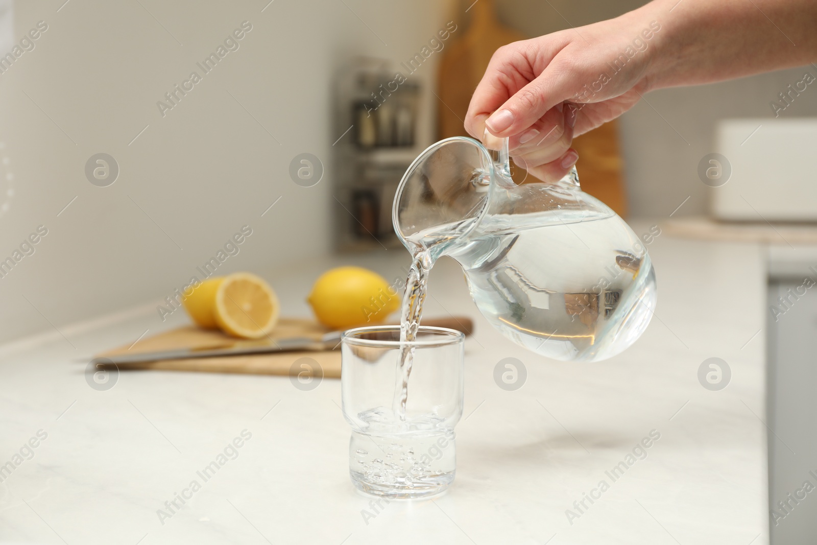 Photo of Woman pouring water from jug into glass at white table in kitchen, closeup