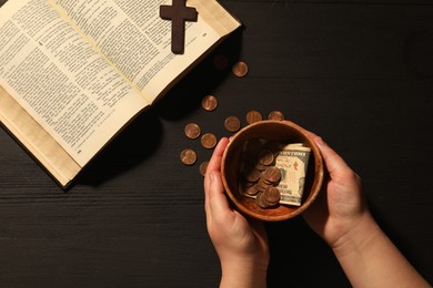 Donate and give concept. Woman with bowl of money, closeup. Bible and cross on black wooden table, top view