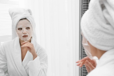 Beautiful woman with sheet mask on her face at mirror indoors