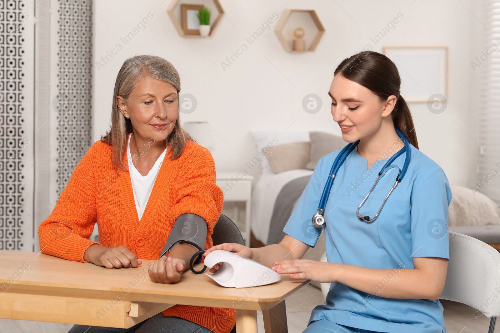 Photo of Young healthcare worker measuring senior woman's blood pressure at wooden table indoors