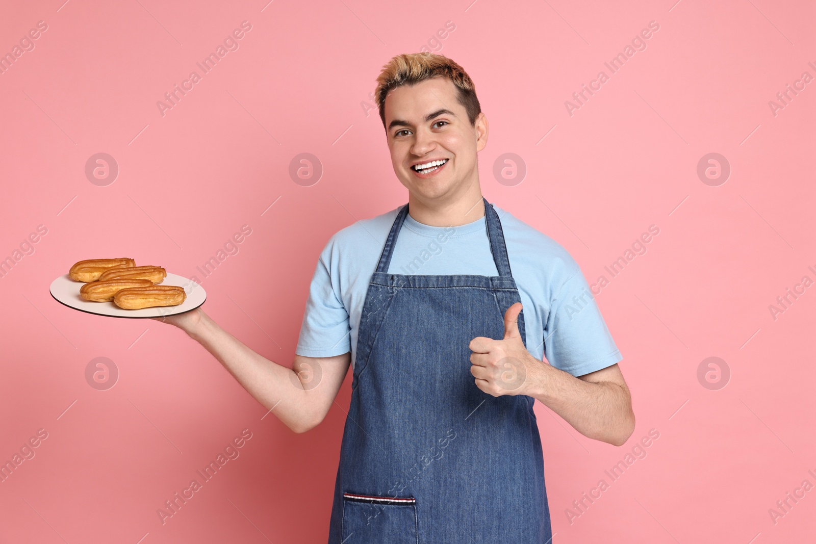 Photo of Portrait of happy confectioner with plate of delicious eclairs showing thumbs up on pink background