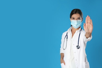 Photo of Doctor in protective mask showing stop gesture on blue background, space for text. Prevent spreading of coronavirus