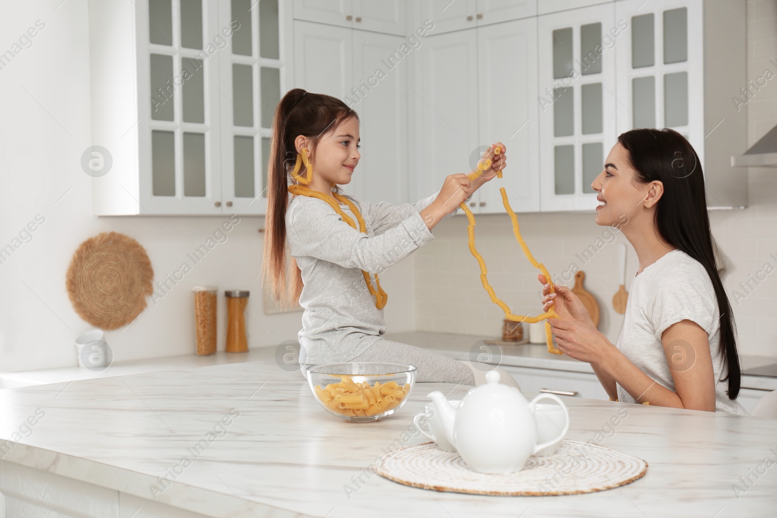 Photo of Young mother and her daughter with necklaces made of pasta having fun in kitchen