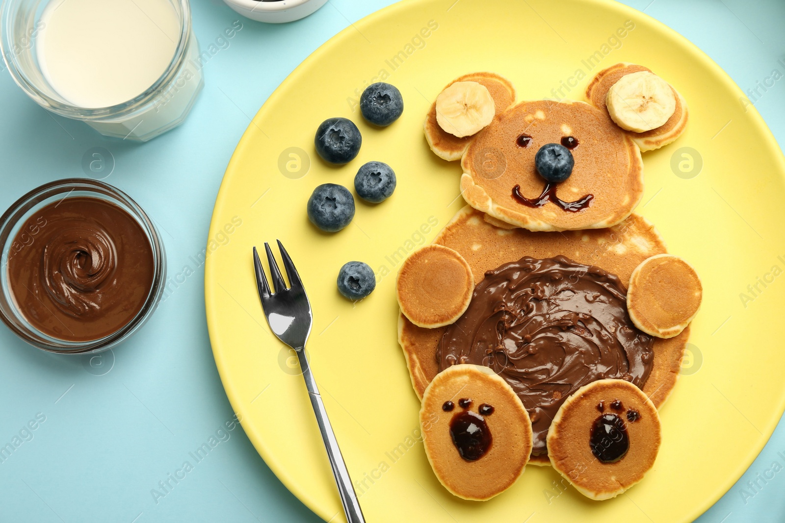 Photo of Creative serving for kids. Plate with cute bear made of pancakes, blueberries, bananas and chocolate paste on light blue table, flat lay