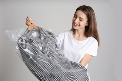 Young woman holding hanger with jacket in plastic bag on grey background. Dry-cleaning service