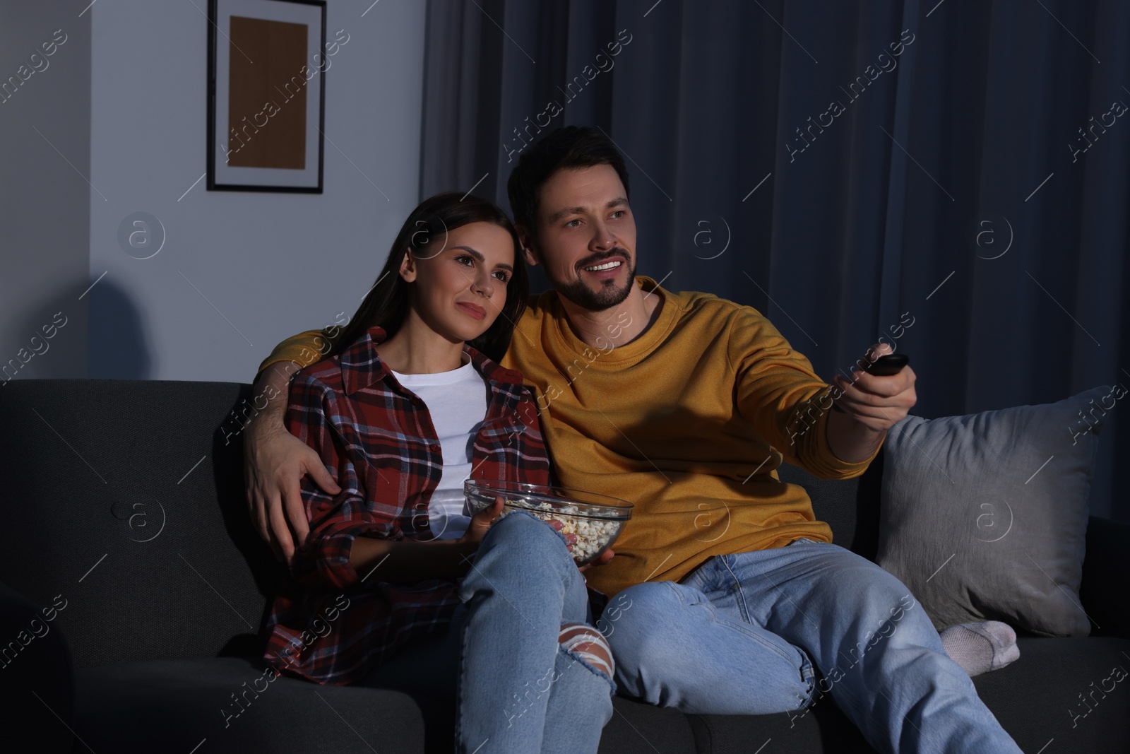 Photo of Couple watching TV at home in evening. Man changing channels with remote control