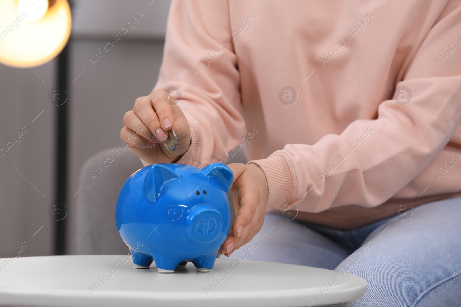 Photo of Young woman putting coin into piggy bank at table indoors, closeup