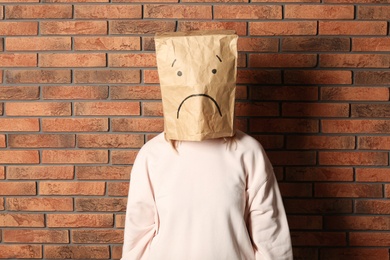 Photo of Woman wearing paper bag with drawn sad face near brick wall