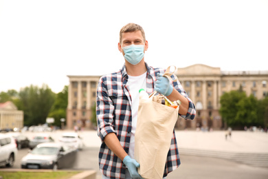 Photo of Male volunteer in protective mask and gloves with products on city street. Aid during coronavirus quarantine