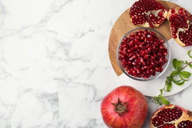 Ripe juicy pomegranate grains and green leaves on white marble table, flat lay. Space for text