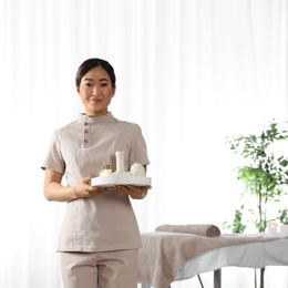 Portrait of young Asian masseuse holding tray with spa stuff in salon, space for text