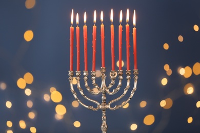Photo of Silver menorah with burning candles against blue background and blurred festive lights. Hanukkah celebration