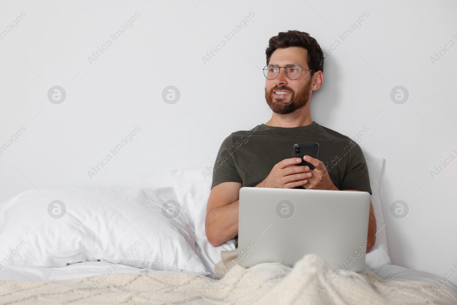 Photo of Handsome man using smartphone and laptop in bed, space for text