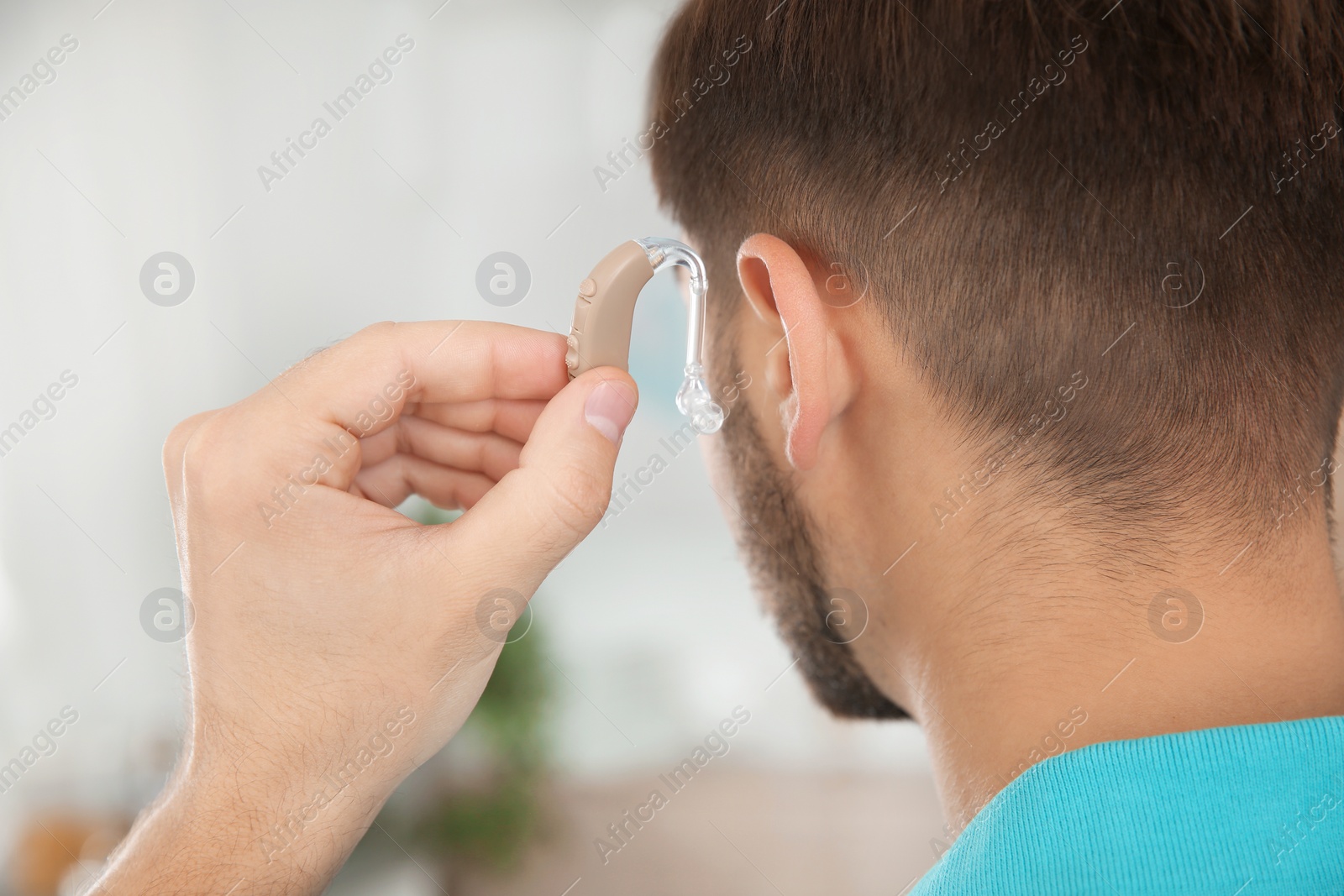 Photo of Young man putting hearing aid in ear indoors, closeup