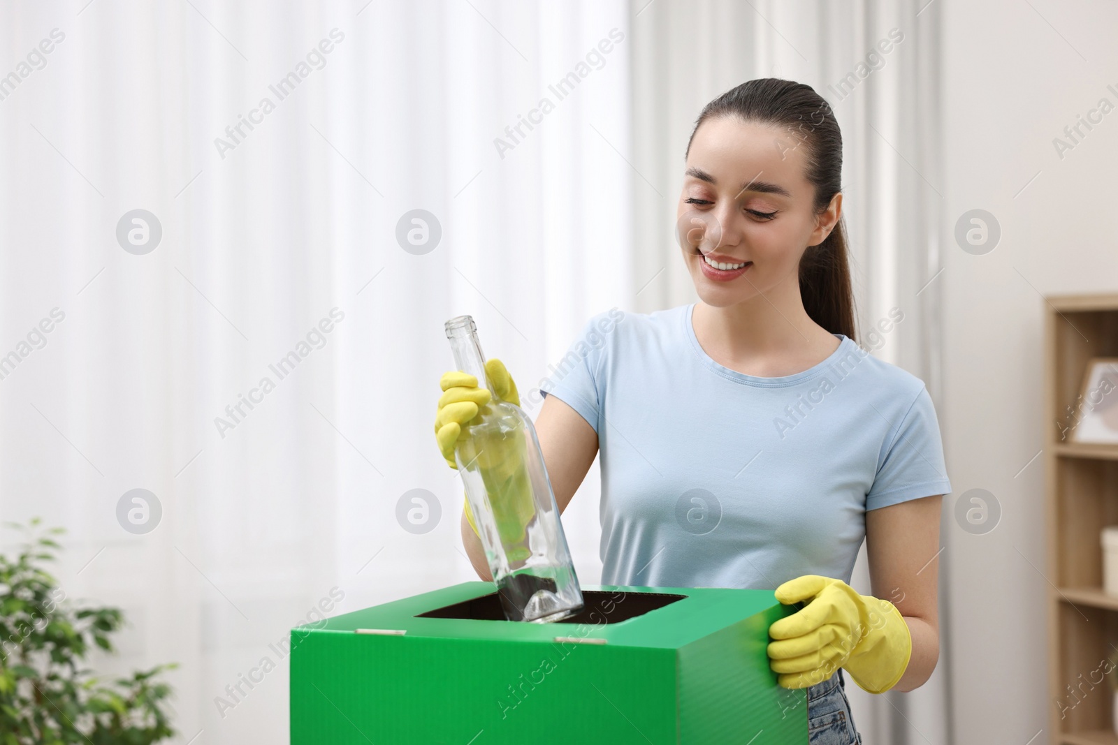 Photo of Garbage sorting. Smiling woman throwing glass bottle into cardboard box in room. Space for text