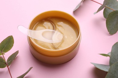 Photo of Under eye patches in jar with spatula and green twigs on light pink background. Cosmetic product