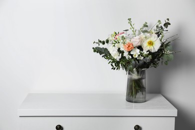 Photo of Bouquet with beautiful flowers in vase on chest of drawers near white wall. Space for text