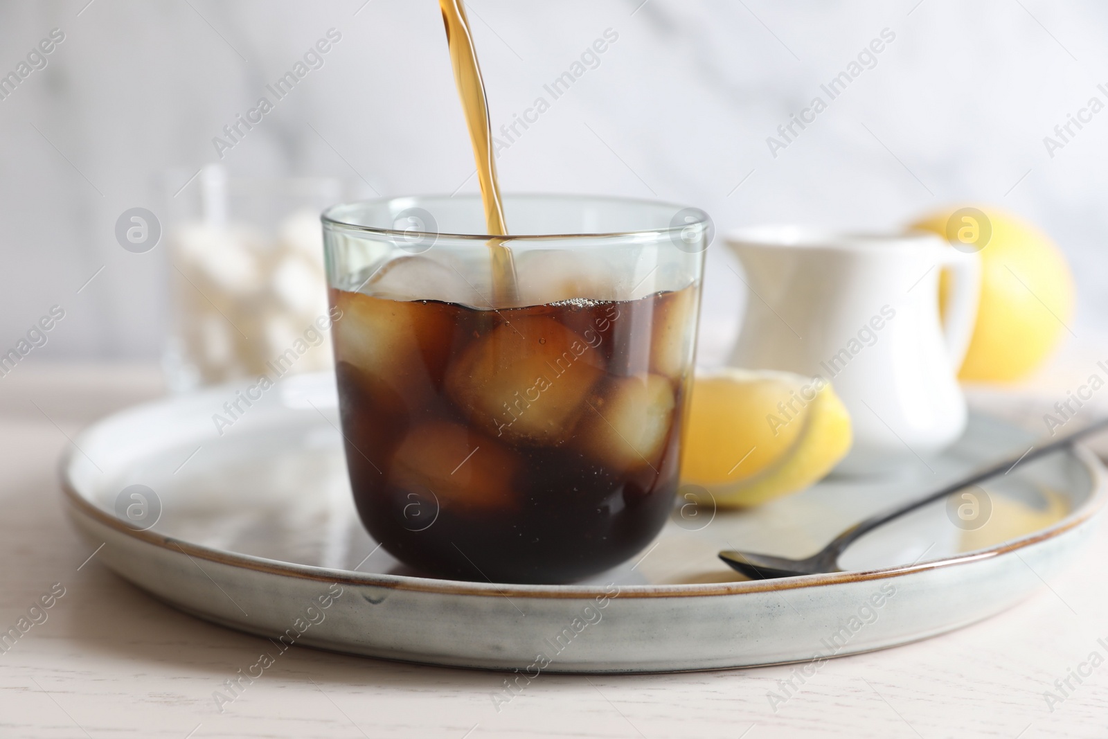 Photo of Pouring coffee into glass with ice cubes at white wooden table, closeup