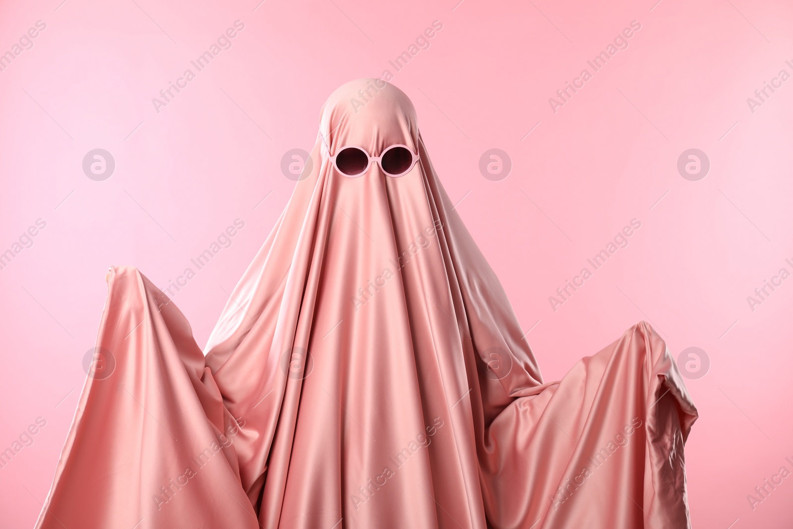 Photo of Glamorous ghost. Woman in sheet with sunglasses on pink background