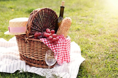 Photo of Wicker basket with bottle of wine, bread, grapes, straw hat, napkin, glass and blanket on green grass outdoors, space for text. Picnic season