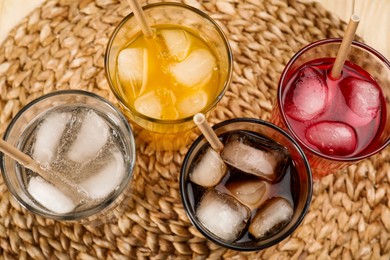 Glasses of different refreshing soda water with ice cubes and straws on wicker mat, flat lay