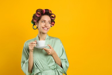 Photo of Beautiful young woman in silk bathrobe with hair curlers and cup of drink on yellow background. Space for text