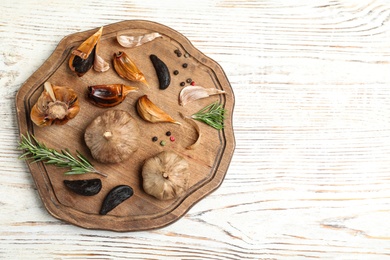 Photo of Board with black garlic and rosemary on white wooden table, top view. Space for text