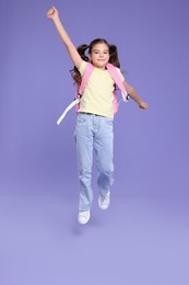 Photo of Cute schoolgirl with backpack jumping on violet background, space for text