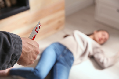 Photo of Man with bloody knife and his victim on floor indoors, closeup. Dangerous criminal