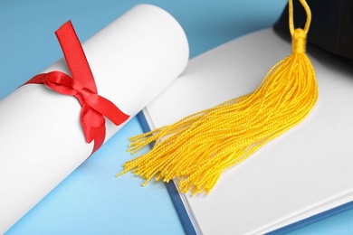 Graduation hat, open book and student's diploma on light blue background, closeup