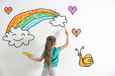 Image of Cute child drawing rainbow and clouds on white wall indoors