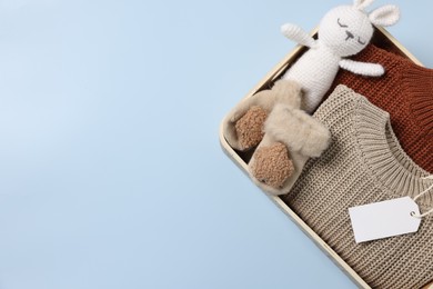Photo of Different baby accessories, knitted sweaters and blank card in box on light blue background, top view. Space for text