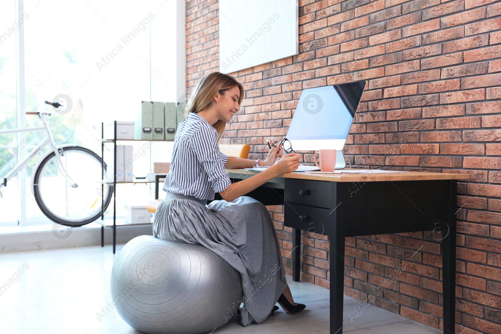 Photo of Beautiful businesswoman sitting on fitness ball at desk in office. Workplace exercises