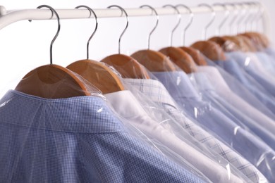 Hangers with shirts in dry cleaning plastic bags on rack against light background, closeup