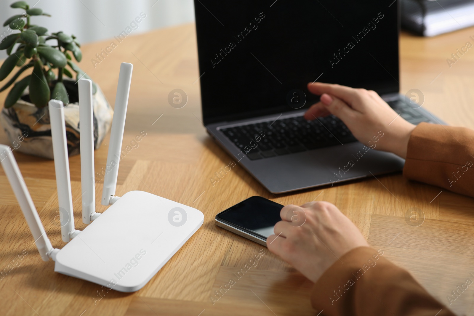 Photo of Woman with smartphone and laptop connecting to internet via Wi-Fi router at table indoors, closeup