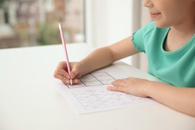 Photo of Little girl solving sudoku puzzle at white table indoors, closeup