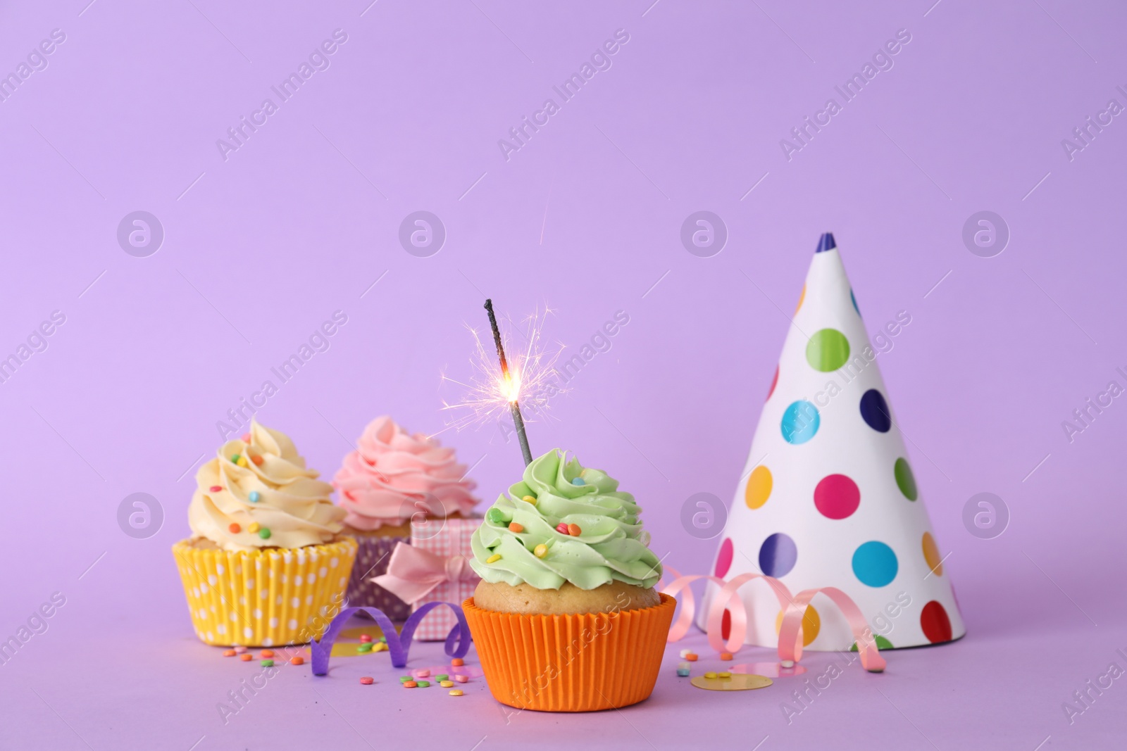 Photo of Birthday cupcakes with burning sparkler, party hat and gift box on violet background